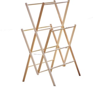 May include: A natural wood drying rack with a folding, X-frame design. The rack has multiple horizontal bars for hanging clothes and is made of light-colored wood. The rack is designed to be collapsible for easy storage.