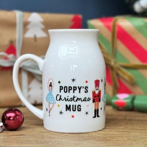 May include: White ceramic mug with a curved handle, featuring the text "POPPY'S Christmas MUG" and illustrations of a ballerina and a nutcracker. The mug has a wide base and a slightly flared top. Christmas gift.