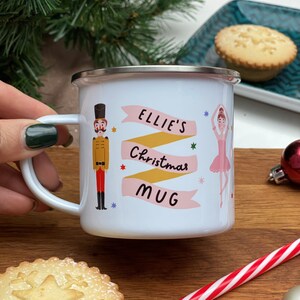 May include: White enamel mug with a pink and yellow banner that says "Ellie's Christmas Mug". The mug features a cartoon illustration of a nutcracker and a ballerina.