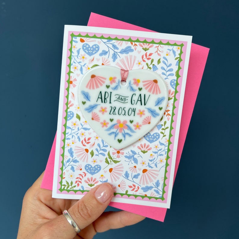 May include: A white ceramic heart with a floral design and the text "ABI and GAV 28.05.04" is attached to a pink and green floral card.