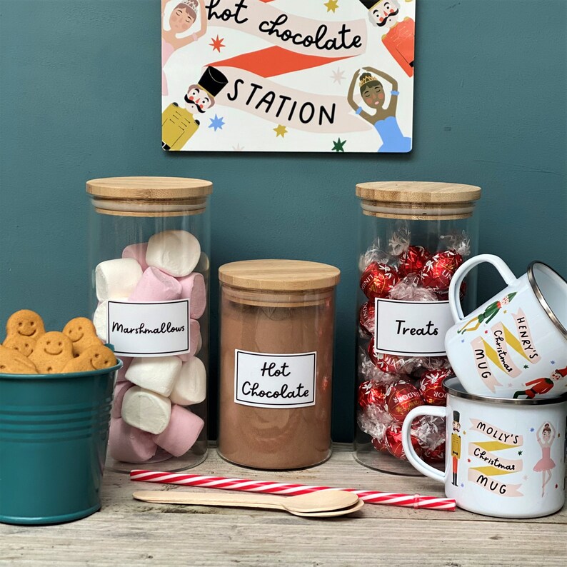 Peut inclure: Trois bocaux en verre avec des couvercles en bambou remplis de guimauves, de chocolat chaud en poudre et de bonbons au chocolat. Les bocaux sont &eacute;tiquet&eacute;s "Marshmallows", "Chocolat chaud" et "Friandises". Deux tasses en &eacute;mail blanc avec des motifs de No&euml;l sont au premier plan. Les tasses sont &eacute;tiquet&eacute;es "Mug de No&euml;l d'Henry" et "Mug de No&euml;l de Molly".