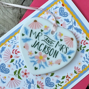 May include: White ceramic heart-shaped ornament with a floral design and the text "Mr. and Mrs. Jackson" in green lettering.