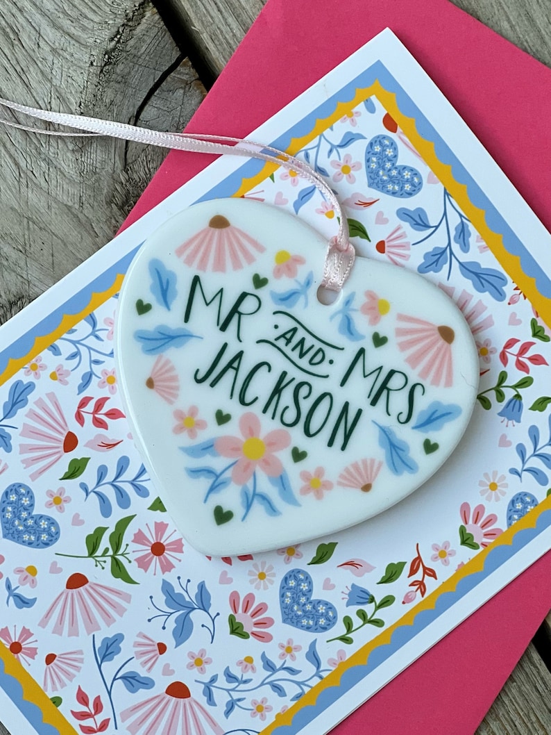 May include: White ceramic heart-shaped ornament with a floral design and the text "Mr. and Mrs. Jackson" in green lettering.