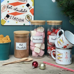 Peut inclure: Trois bocaux en verre avec des couvercles en bambou remplis de m&eacute;lange pour chocolat chaud, de guimauves et de friandises au chocolat. Les bocaux sont &eacute;tiquet&eacute;s "Chocolat chaud", "Guimauves" et "Friandises". Deux tasses en &eacute;mail blanc avec des motifs de No&euml;l et les mots "Mug de No&euml;l de Molly" et "Mug de No&euml;l d'Henry" sont au premier plan.