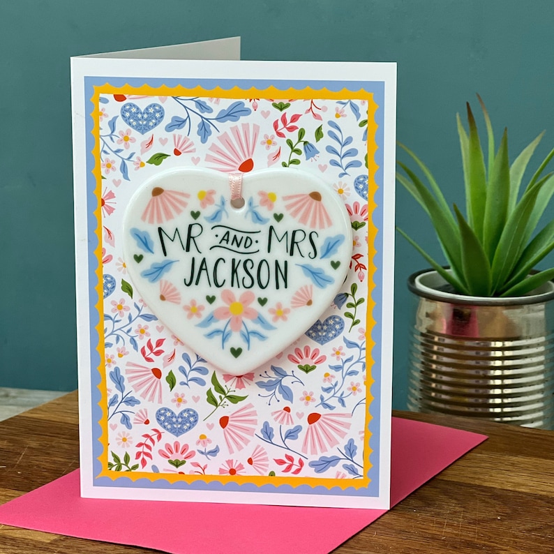 May include: A white ceramic heart-shaped ornament with a floral design and the text "MR. AND MRS. JACKSON" in black lettering. The ornament is attached to a white card with a floral pattern.