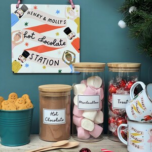 Peut inclure: Trois bocaux en verre avec couvercles, remplis de chocolat chaud, de guimauves et de bonbons rouges et dor&eacute;s. Les bocaux sont &eacute;tiquet&eacute;s "Chocolat chaud", "Guimauves" et "Friandises". Il y a un panneau au-dessus des bocaux qui dit "Henry & Molly's Hot Chocolate Station".