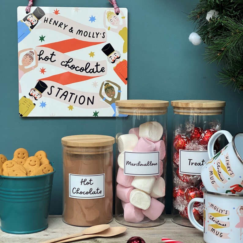Peut inclure: Trois bocaux en verre avec couvercles, remplis de chocolat chaud, de guimauves et de bonbons rouges et dor&eacute;s. Les bocaux sont &eacute;tiquet&eacute;s "Chocolat chaud", "Guimauves" et "Friandises". Il y a un panneau au-dessus des bocaux qui dit "Henry & Molly's Hot Chocolate Station".