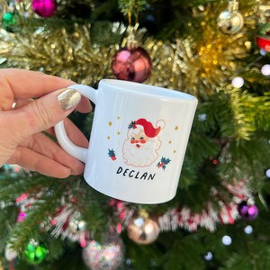 May include: White ceramic mug with a Christmas-themed design featuring a cartoon Santa Claus with a red hat and a white beard. The mug has gold stars and holly berries around the design. The name "DECLAN" is printed in black below the Santa Claus.