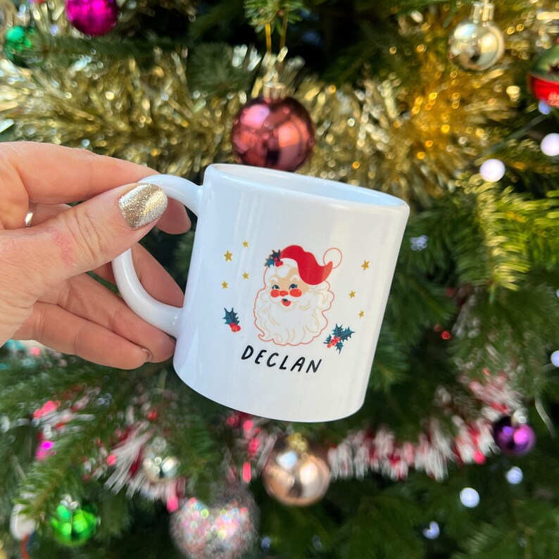 May include: White ceramic mug with a Christmas-themed design featuring a cartoon Santa Claus with a red hat and a white beard. The mug has gold stars and holly berries around the design. The name "DECLAN" is printed in black below the Santa Claus.