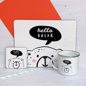 May include: A white ceramic mug with a black and white illustration of a bear with the text "hello ralph" in a speech bubble. The mug has a silver rim and handle. There is a white coaster with a similar design and a white placemat with a larger version of the same design.