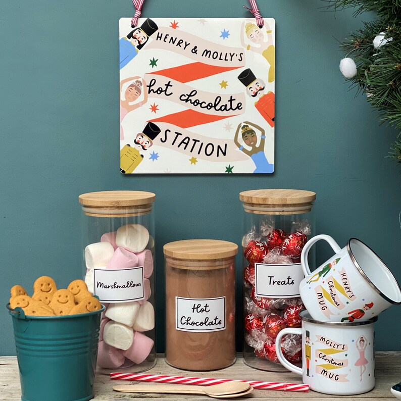 Peut inclure: Un panneau avec le texte "Henry & Molly's Hot Chocolate Station" sur un th&egrave;me festif de Casse-Noisette et de ballerine. Trois bocaux en verre avec des couvercles en bambou contiennent des guimauves, du chocolat chaud en poudre et des friandises au chocolat. Deux tasses en &eacute;mail avec des motifs de No&euml;l et les noms "Henry's" et "Molly's" sont sur une surface en bois.