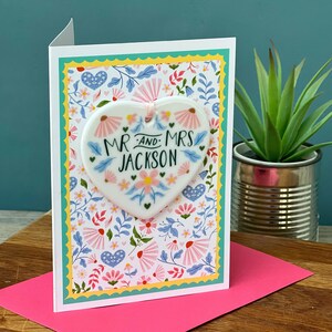 May include: A white ceramic heart-shaped ornament with the text "Mr. and Mrs. Jackson" in black script surrounded by pink and blue flowers. The ornament is attached to a white card with a floral pattern.