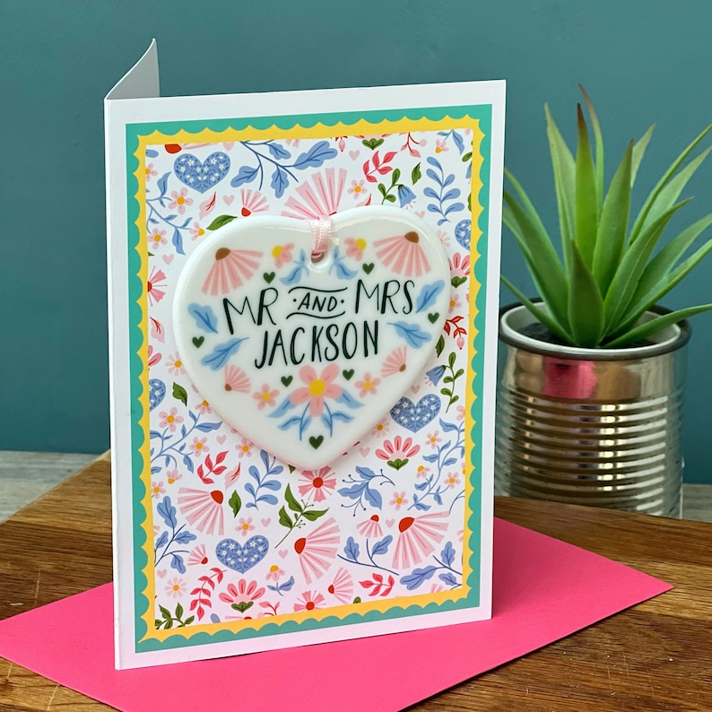 May include: A white ceramic heart-shaped ornament with the text "Mr. and Mrs. Jackson" in black script surrounded by pink and blue flowers. The ornament is attached to a white card with a floral pattern.