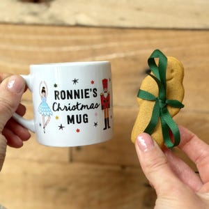 May include: White ceramic mug with a Christmas theme. The mug has a ballerina, a nutcracker, and stars printed on it. The text on the mug reads "Ronnie's Christmas Mug".