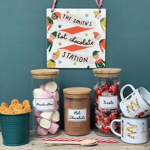 Peut inclure: Un stand de chocolat chaud festif avec trois bocaux en verre remplis de guimauves, de chocolat et de friandises. Les bocaux ont des couvercles en bois et des &eacute;tiquettes qui disent "Marshmallows", "Chocolat chaud" et "Friandises". Il y a aussi une pancarte qui dit "The Smith's Hot Chocolate Station" avec un design d'elfe fantaisiste. Deux tasses avec des motifs de No&euml;l sont &eacute;galement incluses.