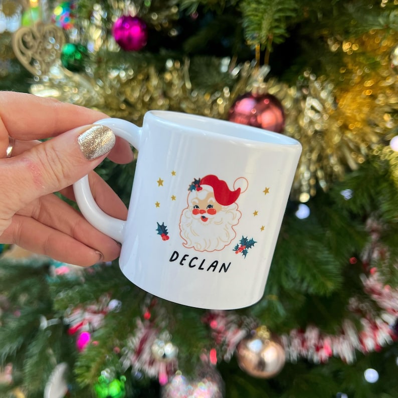 May include: White ceramic mug with a cartoon Santa Claus illustration and the name "Declan" printed on the front. The mug is decorated with gold stars and holly.