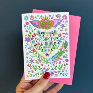 May include: A wedding card with a bright floral design and a pink envelope. The card features a heart-shaped ornament with the text "MR AND MRS WHEELER 19.07.2025". The design includes colourful flowers, birds, and a winged heart.