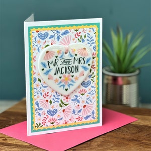 May include: A white ceramic heart ornament with a floral design and the text "Mr. and Mrs. Jackson" on a card with a floral pattern.