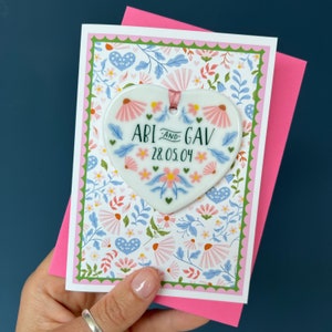 May include: A white heart-shaped ceramic ornament with a floral design and the text "Abi and Gav 28.05.04" on a pink and white floral card.