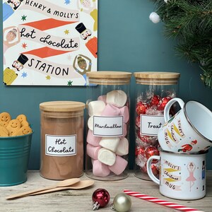 Peut inclure: Un ensemble de quatre bocaux en verre avec des couvercles en bambou, remplis de m&eacute;lange pour chocolat chaud, de guimauves et de bonbons. Les bocaux sont &eacute;tiquet&eacute;s "Chocolat chaud", "Guimauves" et "Friandises". Il y a deux tasses avec des motifs de No&euml;l et les noms "Mug de No&euml;l d'Henry" et "Mug de No&euml;l de Molly".
