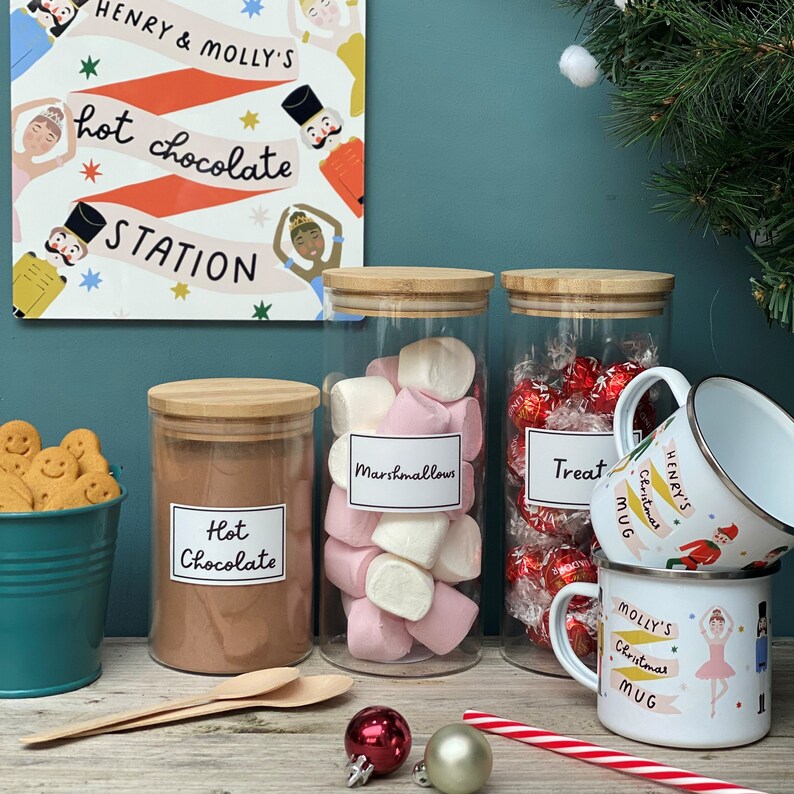 Peut inclure: Un ensemble de quatre bocaux en verre avec des couvercles en bambou, remplis de m&eacute;lange pour chocolat chaud, de guimauves et de bonbons. Les bocaux sont &eacute;tiquet&eacute;s "Chocolat chaud", "Guimauves" et "Friandises". Il y a deux tasses avec des motifs de No&euml;l et les noms "Mug de No&euml;l d'Henry" et "Mug de No&euml;l de Molly".