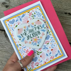 May include: A pink card with a white heart-shaped ornament attached. The ornament has a floral design and the text "Mr. and Mrs. Jackson". The card has a floral pattern with blue, yellow, and pink flowers.
