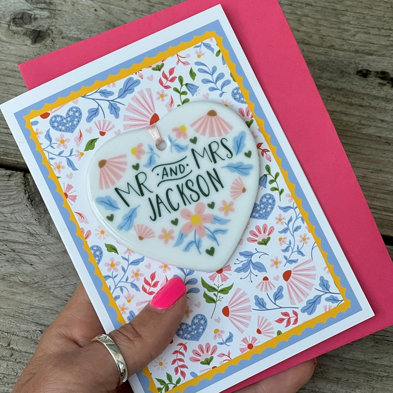 May include: A pink card with a white heart-shaped ornament attached. The ornament has a floral design and the text "Mr. and Mrs. Jackson". The card has a floral pattern with blue, yellow, and pink flowers.