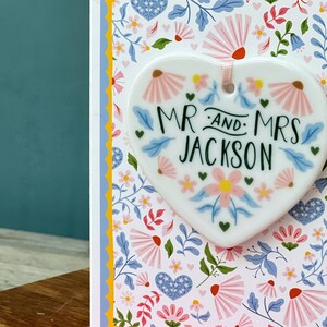 May include: White ceramic heart-shaped ornament with a floral design and the text "Mr. and Mrs. Jackson" in green lettering.