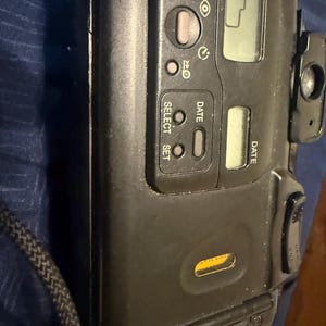 K&ouml;nnte beinhalten: Eine schwarze Point-and-Shoot-Filmkamera mit digitaler Anzeige und mehreren Tasten. Die Kamera hat einen eingebauten Blitz und ein Objektiv an der Seite. Die Kamera hat einen schwarzen Riemen.
