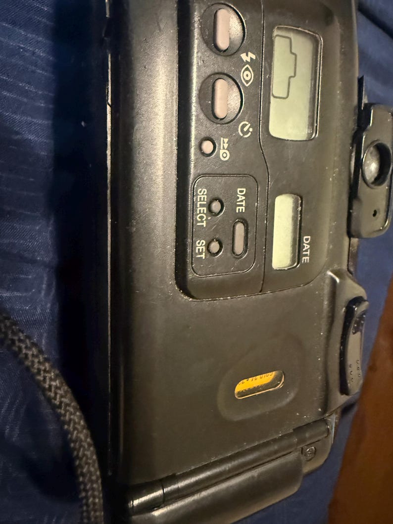 K&ouml;nnte beinhalten: Eine schwarze Point-and-Shoot-Filmkamera mit digitaler Anzeige und mehreren Tasten. Die Kamera hat einen eingebauten Blitz und ein Objektiv an der Seite. Die Kamera hat einen schwarzen Riemen.