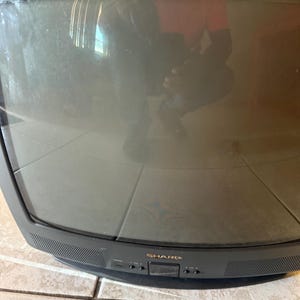 Puede incluir: Un televisor Sharp negro con una pantalla curva. La pantalla refleja el entorno circundante. La parte inferior del televisor tiene botones y el nombre de la marca "SHARP".