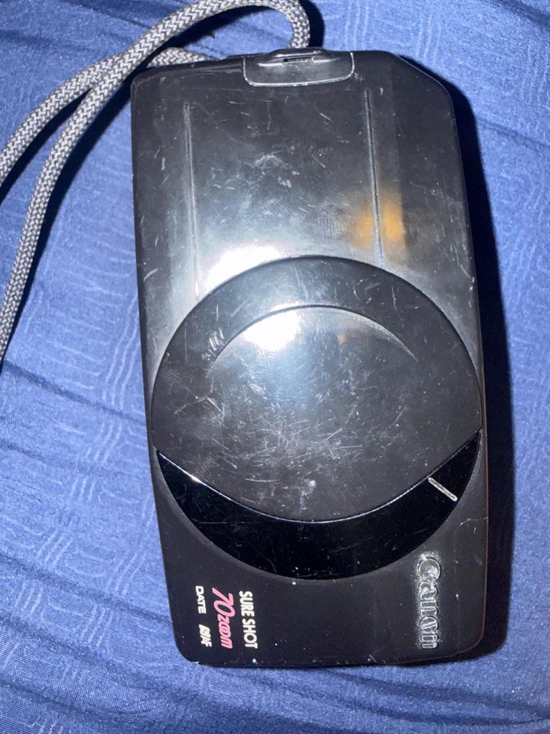 K&ouml;nnte beinhalten: Schwarze Canon Sure Shot 70 Zoom Kamera mit grauem Gurt. Die Kamera hat ein gro&szlig;es Objektiv und die W&ouml;rter "SURE SHOT 70 Zoom" und "DATE AOF" sind auf der Vorderseite aufgedruckt. Die Kamera ist rechteckig mit abgerundeten Kanten.