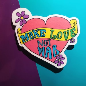 Könnte beinhalten: Ein rosa herzförmiger Aufkleber mit einem gelben Banner, auf dem "Make Love Not War" steht. Das Banner ist grün umrandet. Der Aufkleber hat lila Blumen an den Rändern.