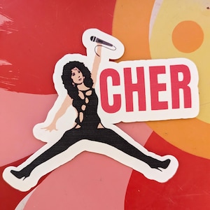 May include: A white sticker with a black and white illustration of Cher in a black jumpsuit holding a microphone. The word "CHER" is in red letters above her.