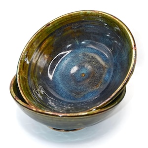 Puede incluir: Dos cuencos de cerámica apilados. Los cuencos tienen un interior azul oscuro con un acabado brillante. Los bordes son de color verde oscuro con un borde dorado. El exterior de los cuencos es de color verde oscuro.