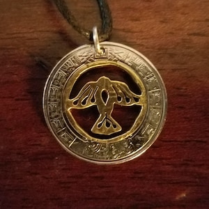 May include: A silver coin necklace with a gold bird cut-out pendant. The coin has a circular design with a repeating pattern around the edge.
