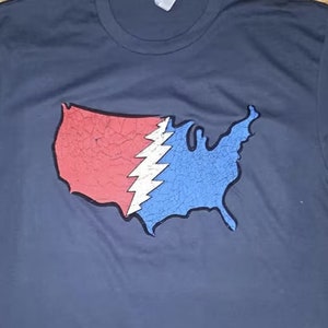 May include: Navy blue t-shirt featuring a distressed graphic of the United States map divided into red and blue sections, with a white lightning bolt design in the center. The shirt is made of a soft material.
