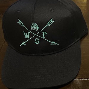 Peut inclure: Une casquette de baseball noire avec un motif brodé vert de deux flèches croisées avec une coiffe amérindienne au centre. Les lettres WSP sont brodées au centre du motif.