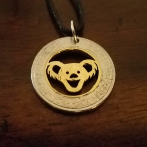 May include: A silver and gold pendant necklace featuring a bear design. The pendant has a circular shape with a gold bear cutout in the center, surrounded by a silver border. The necklace has a black cord.