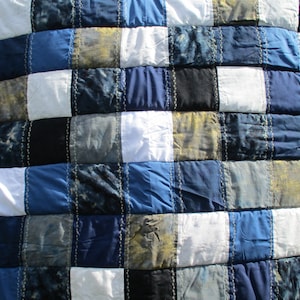 May include: A patchwork quilt in shades of blue, white, black, and gold. The squares are stitched together with white thread. The quilt's design is a grid of squares. The quilt is likely made of cotton or a similar fabric. The quilt is a handmade item.