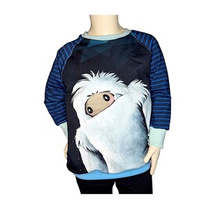 Könnte beinhalten: Ein schwarzes Sweatshirt mit blau-weiß gestreiften Ärmeln. Die Vorderseite des Sweatshirts zeigt ein weißes, flauschiges Wesen mit einem braunen Gesicht, das hinter seinem Fell hervorlugt.