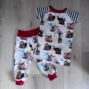 Könnte beinhalten: Ein weißer Baby-Strampler mit rotem Besatz und einem blau-weiß gestreiften Ärmel. Der Strampler hat ein Muster aus Tieren mit bunten Farbflecken. Der Strampler wird mit passenden roten Hosen kombiniert.