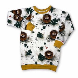 Könnte beinhalten: Weißes Langarm-Sweatshirt mit einem sich wiederholenden Muster aus Cartoon-Löwen, grünen Blättern und roten Herzen. Die Bündchen und der Halsausschnitt sind senfgelb. Das Sweatshirt hat einen Rundhalsausschnitt und eine lockere Passform.