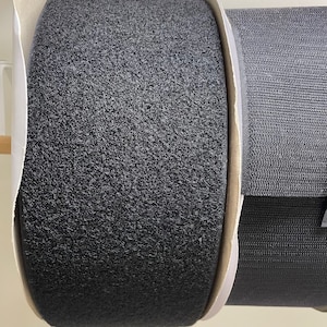 Puede incluir: Dos rollos de cinta de velcro. Un rollo es de color gris oscuro, con una textura difusa, y el otro es de color gris más oscuro, con una textura tejida. Ambos rollos están sobre núcleos de cartón con bordes de papel blanco.