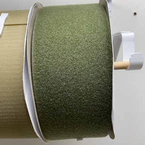 Puede incluir: Primer plano de dos rollos de tela. Uno es verde oliva, el otro beige. Están montados en un soporte metálico blanco con una clavija de madera. La tela tiene una superficie texturizada y difusa. Ideal para manualidades o tapicería.