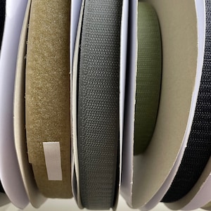 May include: Close-up of four rolls of hook and loop fastener tape. The rolls are in different colors: beige, black, green, and black. The tape is used for attaching two pieces of fabric together.
