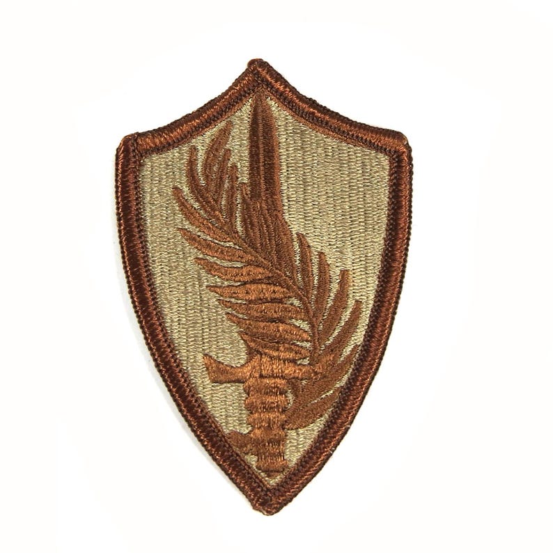 May include: A brown embroidered patch with a tan background. The patch is shaped like a shield and features a stylized brown leaf with a sword through it.