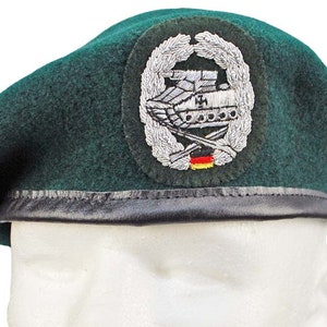 Puede incluir: Un boina verde oscuro con una banda de cuero negro. La boina tiene un parche bordado en plata con un tanque, rifles cruzados y una bandera alemana.