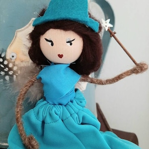 May include: A handmade doll with brown hair, wearing a turquoise dress and hat, holding a twig with a white star on the end.