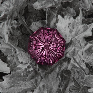 May include: A faceted, octagonal, magenta-colored glass object. The object has a starburst design emanating from the center. The background is a grayscale image of leaves.